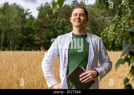Smiling farmer standing confidently in wheat field holding tablet and folder near forest edge. Successful agriculture concept showing pride in product Stock Photo