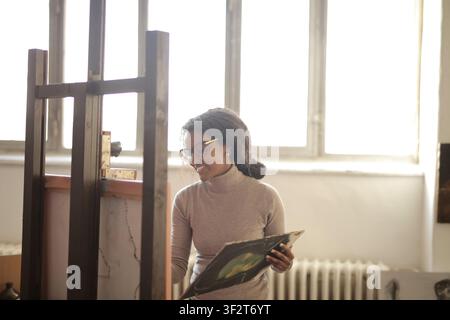 Smiling artist stands by easel in bright studio Stock Photo