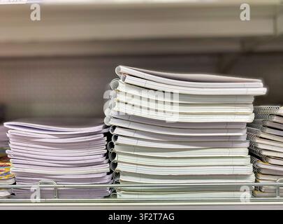 Neatly arranged stack of white-covered notebooks with spiral bindings on a shelf in a stationery store, with colorful items visible in the background Stock Photo