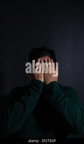 Conceptual portrait of a distressed Asian male wearing a dark green long sleeve shirt covering both eyes with hands against a solid black background. Stock Photo