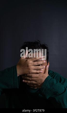 Striking portrait of an Asian male wearing a dark green shirt peeking through fingers while covering the face against a solid black background. The su Stock Photo