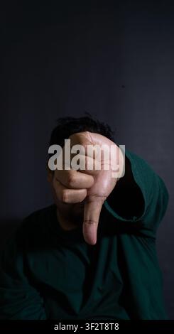 Conceptual studio photograph focusing on a hand making a thumbs down gesture with a blurred Indonesian man in the stark black background. The downward Stock Photo