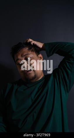 Conceptual portrait of a confused adult Indonesian male wearing a dark green shirt scratching the head against a solid black background. The individua Stock Photo