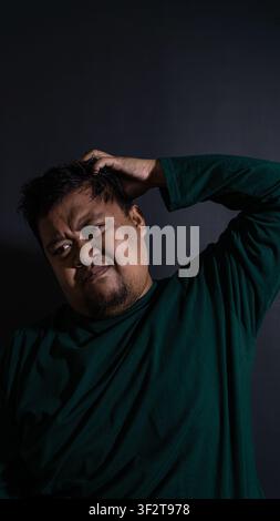 Humorous yet moody portrait of an uncertain adult Indonesian male wearing a dark green shirt scratching the head against a deep black background. The Stock Photo