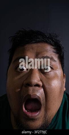 Extreme close up portrait of an adult Indonesian male wearing a dark green shirt shouting with a wide open mouth against a black background. The subje Stock Photo