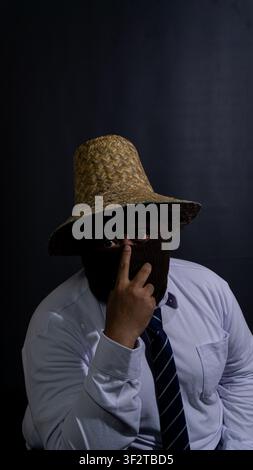 Adult Southeast Asian male wearing a brown knit balaclava mask and a traditional woven straw hat while pointing a single finger directly at the camera Stock Photo