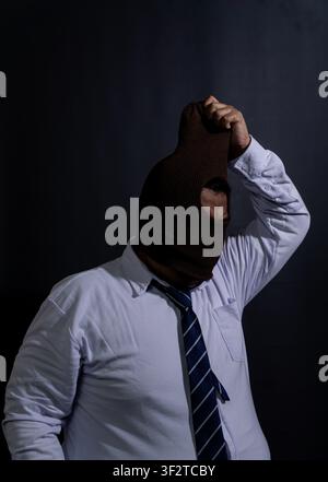 Adult Southeast Asian male caught in the act of pulling a brown knit balaclava over the head against a dark black background. The businessman wears a Stock Photo