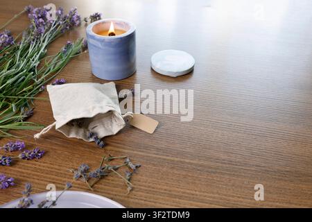 Open cotton sachet filled with dried lavender flowers on table. Scented candle burning near fabric pouch with blank craft tag. Cosy aromatherapy Stock Photo