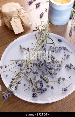 Dried lavender flowers on white ceramic plate top view. Small honey jar with paper tag near concrete candle container. Botanical herbarium workspace Stock Photo