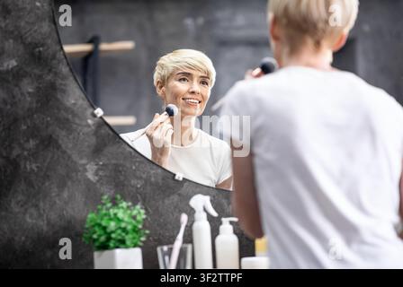 Mature Lady Using Cosmetic Brush Applying Face Powder In Bathroom Stock Photo