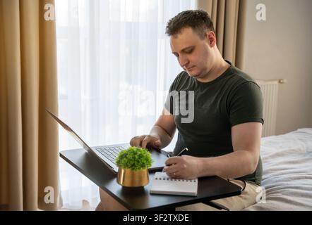 35 year old Caucasian man sits at a table, works on a laptop, and writes in a notebook in his apartment bedroom. Remote work and freelance concept. Fo Stock Photo