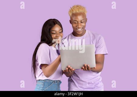 Happy young black couple with laptop browsing web, chatting with friends, watching cool video on lilac studio background Stock Photo