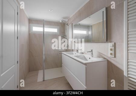Modern bathroom interior with double vanity, large wall mirror, frameless glass shower, chrome fixtures, beige tile, bright natural light, clean Stock Photo
