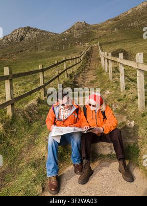 Young male & senior female hiker wearing orange jackets sitting on pathway up steep hill studying map Stock Photo