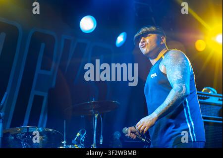 Tattooed lead vocalist wearing a flat cap performing live on stage with heavy metal band Crashout. Stock Photo