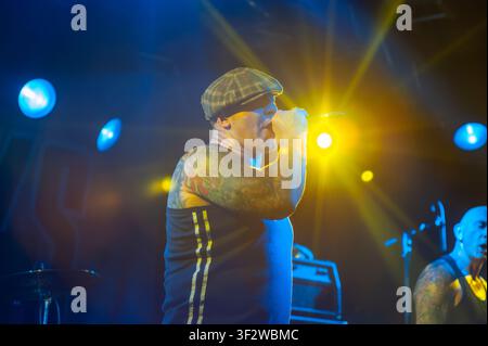 Tattooed lead vocalist wearing a flat cap performing live on stage with heavy metal band Crashout. Stock Photo