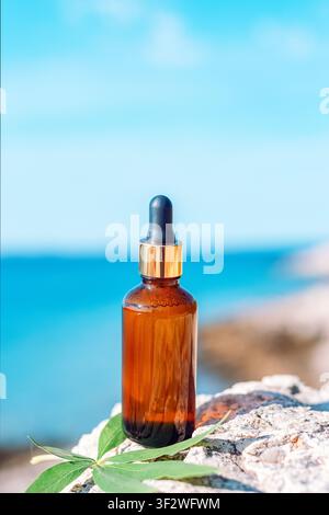 Amber glass dropper bottle with cosmetic serum on seaside rocks with green leaves and blurred blue ocean in natural sunlight. Copy space. Stock Photo