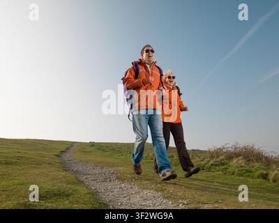 Young male & senior female hikers laughing as they run downhill wearing orange jackets Stock Photo