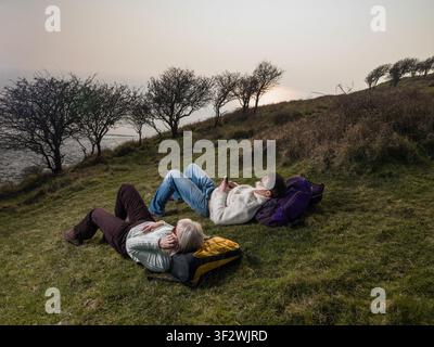 View of young male & senior female hikers laying on backpacks using mobile phones in coastal field Stock Photo
