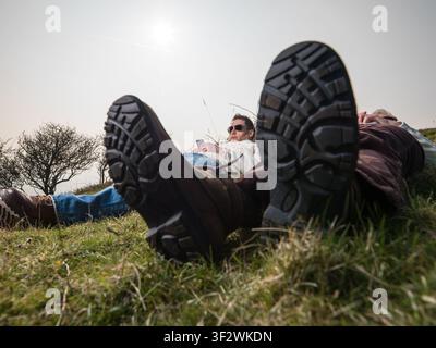 Young male & senior female hikers laying in field using moblie phones at sunset low camera angle view with soles of boots to camera Stock Photo