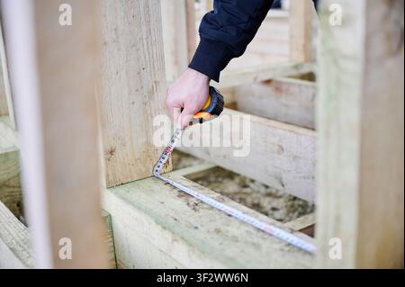 Close up of builder using measuring tape on timber frame corner, verifying width and precise placement of wooden elements during construction work Stock Photo