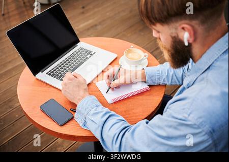 Top view of man writing in notebook beside laptop, coffee cup, and smartphone at outdoor terrace table, showing focused remote work and planning routine Stock Photo
