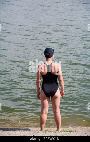 Woman standing in lake preparing for open water swimming. Female swimmer in black swimsuit and cap facing water, back view, outdoor training, summer activity Stock Photo