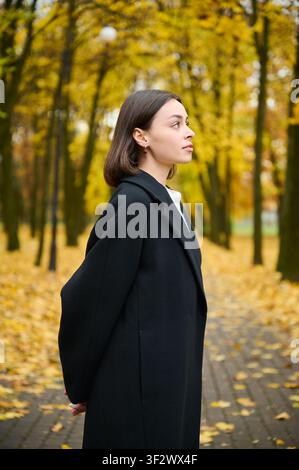 Stylish young woman posing outdoors in fall, dressed in black coat among golden trees and fallen leaves. Perfect for apparel promotion, beauty campaigns, and autumn marketing Stock Photo