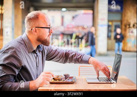 Man using laptop while eating at outdoor cafe, focused freelancer working during lunch break, urban remote work lifestyle and multitasking concept Stock Photo