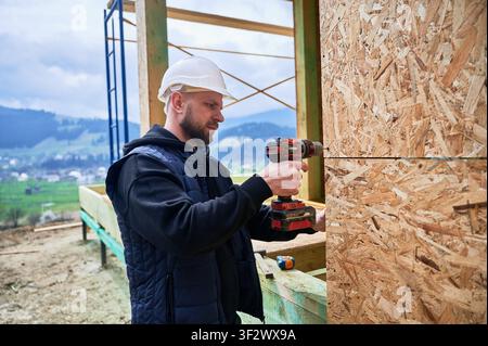 Construction worker installing OSB panels on wooden frame using cordless drill, representing wall assembly, home building process and structural development Stock Photo