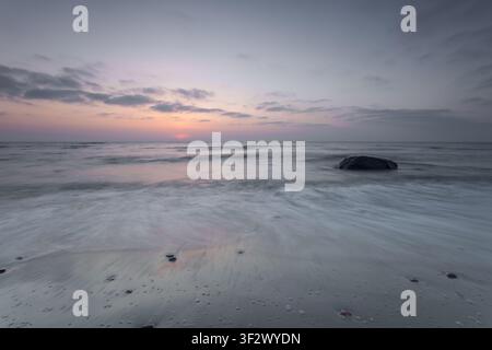 Calm sea at sunrise with soft pastel sky, gentle waves and a single rock in the water, captured with long exposure for a peaceful and dreamy coastal s Stock Photo