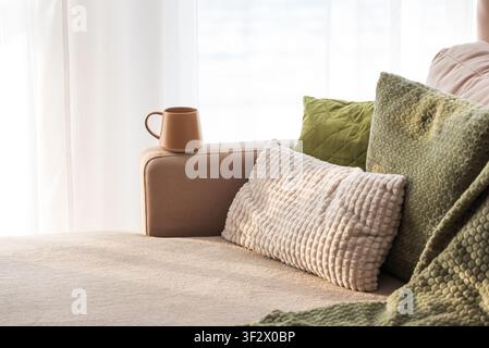 Living room sofa features warm coffee mug placed on its armrest, inviting comfort and relaxation with various plush pillows and a soft blanket near a Stock Photo