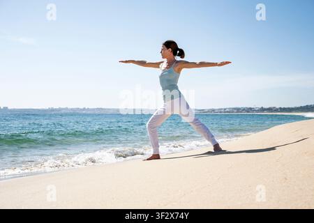 woman practices yoga in Warrior II pose on a sunny sandy beach Stock Photo