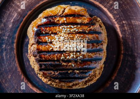Golden brown homemade pastry topped with sesame seeds and egg wash glaze on rustic clay plate, close up. Freshly baked artisan bun captured from above Stock Photo