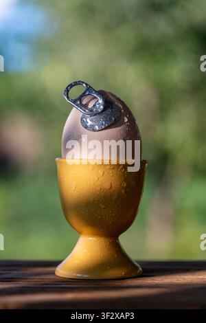 Conceptual egg with aluminum pull tab in holder outdoor, close up. Creative concept of fresh egg with an easy open metal can tab in yellow ceramic cup Stock Photo
