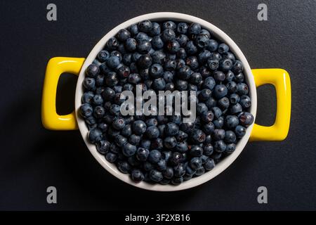 Fresh blueberries in white bowl with yellow handles, close up. Top view of round ceramic pot filled with ripe fresh blueberries on dark background. He Stock Photo