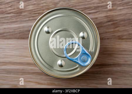 Top view close up of sealed round aluminum tin can with pull tab mechanism, placed on textured brown wooden background. Simple food packaging mockup c Stock Photo