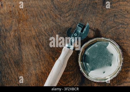 Top view close up of an old retro style can opener with wooden handle next to metal tin lid on dark scratched wooden surface with empty space Stock Photo