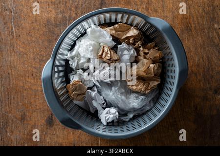 Top view close up of grey plastic garbage bin filled with wrinkled brown and white paper sheets and plastic bag, placed on rustic dark wooden surface. Stock Photo