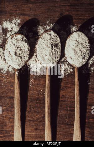 Wooden spoons with white flour on dark background, close up. Three wooden spoons filled with white flour on rustic wooden table with scattered powder Stock Photo