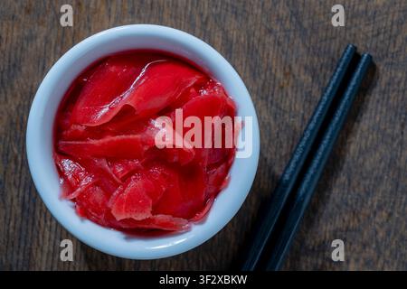 Pickled ginger in white bowl with chopsticks, close up. Top view pink pickled ginger slices in small ceramic bowl next to black chopsticks on wooden t Stock Photo