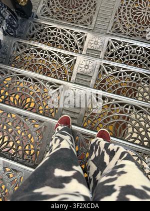 Ornate metal staircase with person wearing red shoes and patterned pants, seen from above. Mobile photo Stock Photo