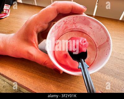 Hand holding a plastic spoon with the last scoop of pink sorbet in a mostly empty paper cup Stock Photo