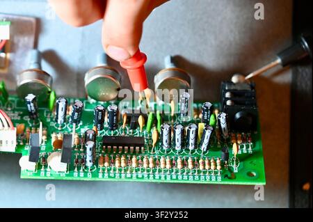 Close-up of a technician testing a green electronic printed circuit board (PCB) with a red multimeter digital probe. Repair, diagnostic, and maintenance process of modern microelectronics and hardware equipment in workshop. Testing Circuit Board with Multimeter Probe Close-up. High quality photo Stock Photo
