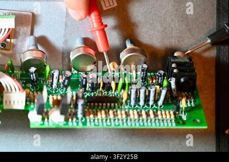 Close-up of a technician testing a green electronic printed circuit board (PCB) with a red multimeter digital probe. Repair, diagnostic, and maintenance process of modern microelectronics and hardware equipment in workshop. Testing Circuit Board with Multimeter Probe Close-up. High quality photo Stock Photo