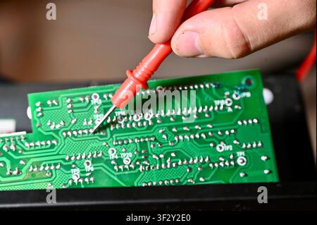 Technician Testing Circuit Board with Multimeter Probe Close-up. High quality Close-up of a technician testing a green electronic printed circuit board (PCB) with a red multimeter digital probe. Repair, diagnostic, and maintenance process of modern microelectronics and hardware equipment in workshop. Stock Photo