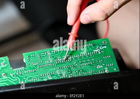 Technician Testing Circuit Board with Multimeter Probe Close-up. High quality Close-up of a technician testing a green electronic printed circuit board (PCB) with a red multimeter digital probe. Repair, diagnostic, and maintenance process of modern microelectronics and hardware equipment in workshop. Stock Photo
