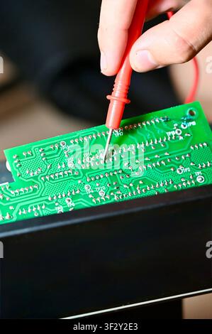 Technician Testing Circuit Board with Multimeter Probe Close-up. High quality Close-up of a technician testing a green electronic printed circuit board (PCB) with a red multimeter digital probe. Repair, diagnostic, and maintenance process of modern microelectronics and hardware equipment in workshop. Stock Photo