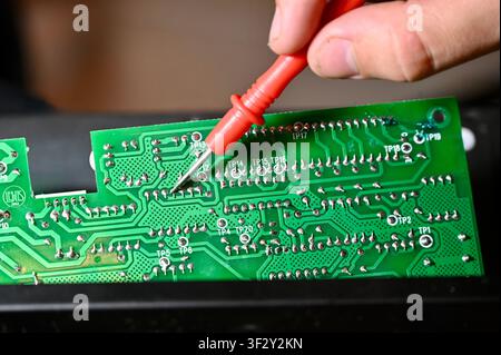 Technician Testing Circuit Board with Multimeter Probe Close-up. High quality Close-up of a technician testing a green electronic printed circuit board (PCB) with a red multimeter digital probe. Repair, diagnostic, and maintenance process of modern microelectronics and hardware equipment in workshop. Stock Photo