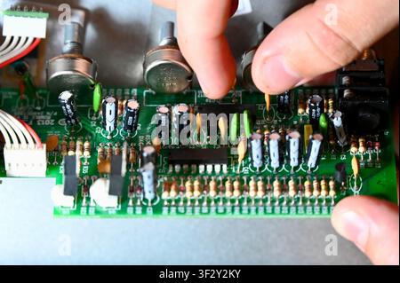 Close-up of an engineer's hands holding a green printed circuit board (PCB) with various electronic components such as capacitors, resistors, potentiometers, and microchips. Concept of microelectronics repair, hardware engineering, DIY audio equipment maintenance, and technology development. Stock Photo